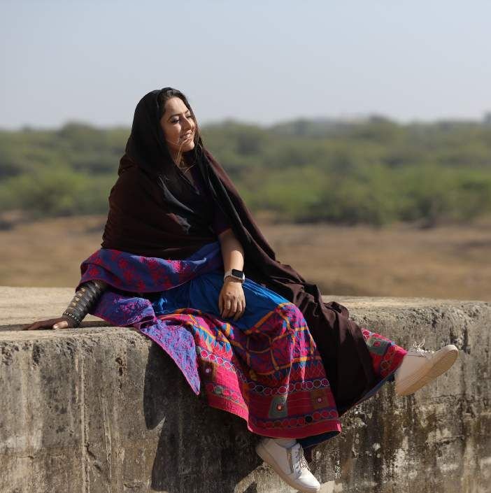 Archie seated on a stone wall in a Kutchi embroidered skirt and dark shawl